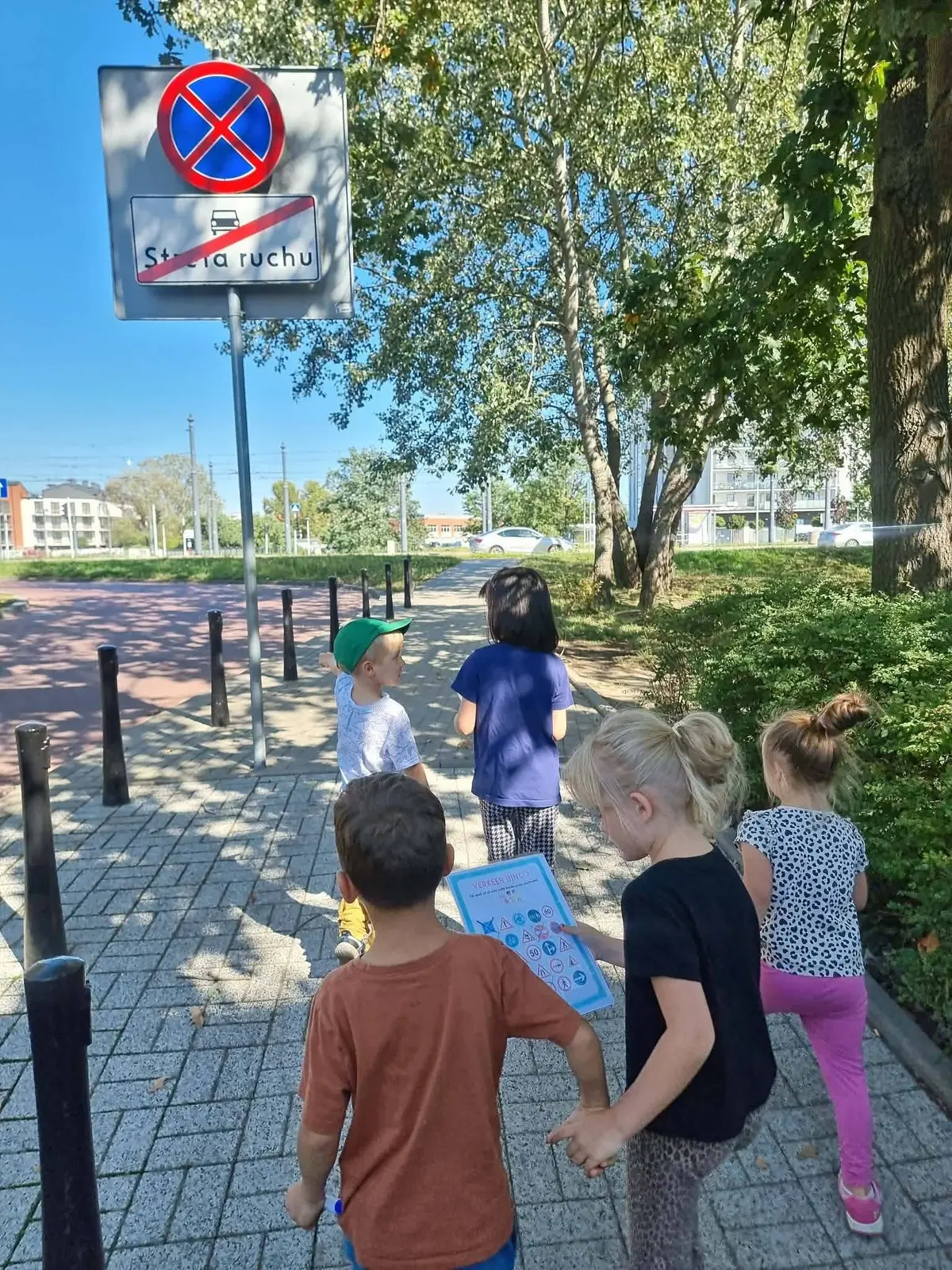 Przedszkolaki z kartami do bingo idą parami chodnikiem obok znaku zakazu zatrzymywania się